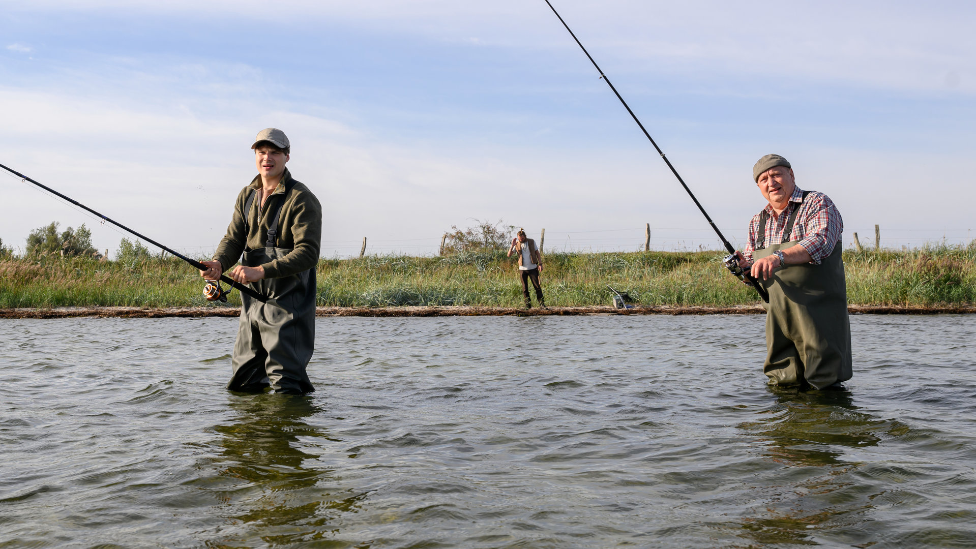 "SOKO Wismar - Grabräuber": Jan-Hinrich Reuter (Udo Kroschwald) und Eddi Jansons (Gustavs Gailus) stehen im Wasser und angeln. Am Ufer steht Gunnar Schmied (Zhenja Isaak).