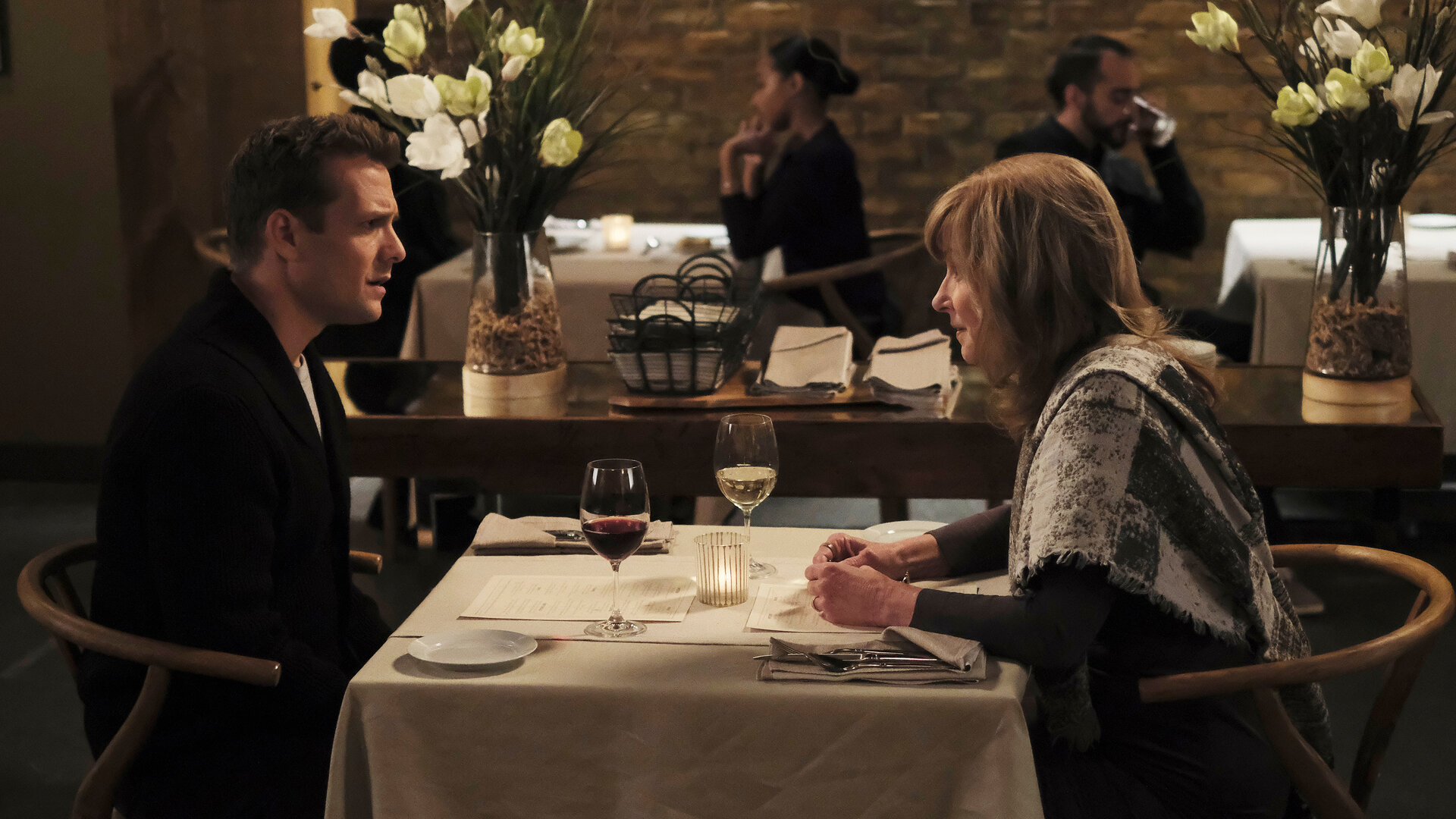 "Suits - Das Gemälde": Harvey (Gabriel Macht) schaut angespannt während er seiner Mutter (Brynn Thayer) in einem Restaurant gegenüber sitzt. Im Hintergrund sitzen weitere Gäste (Komparsen) an Tischen, an der Seite stehen zwei große weiße Blumenarrangements.