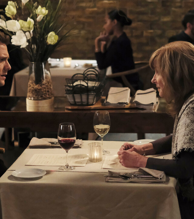 "Suits - Das Gemälde": Harvey (Gabriel Macht) schaut angespannt während er seiner Mutter (Brynn Thayer) in einem Restaurant gegenüber sitzt. Im Hintergrund sitzen weitere Gäste (Komparsen) an Tischen, an der Seite stehen zwei große weiße Blumenarrangements.