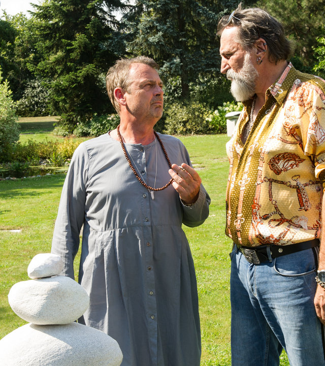 "SOKO Stuttgart - Good Vibrations": Dr. Klas Eckert (Krystian Martinek) steht neben Jack Silber (Andreas Hoppe) im Garten der Entzugsklinik. Während die beiden miteinander sprechen, hält Klas Eckert seine Gebetskette in der Hand. Jack Silber trägt ein gelbes Hemd mit Mustern. Seine Sonnenbrille hat er sich in die Haare geschoben. Klas Eckert trägt eine Art graue Kutte und schaut zu Jack Silber.  Im Hintergrund sieht man ein Blumenbeet, Bäume und eine weiße Statue.