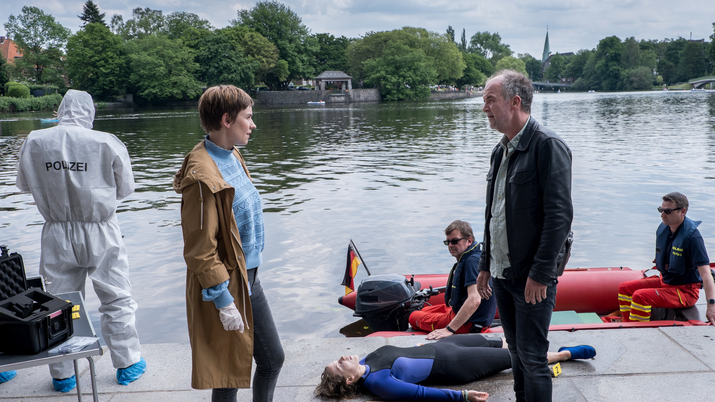"SOKO Hamburg - Trübe Wasser": Oskar Schütz (Marek Erhardt) und Jette Lorenzen (Gundula Niemeyer) stehen sich an einem Tatort am Fluss gegenüber und unterhalten sich. Rechts neben ihnen liegt die tote Jule Neumann (Lara Maria Wichels), die einen blau-schwarzen Wassersportanzug trägt. Auf dem Fluss befinden sich zwei Rettungskräfte (Komparsen) in einem roten Boot, an dem eine kleine Deutschlandfahne steckt. Links im Bild steht mit dem Rücken zur Kamera ein Mitglied der Spurensicherung in einem weißen Overall, auf dem der Begriff "Polizei" steht.