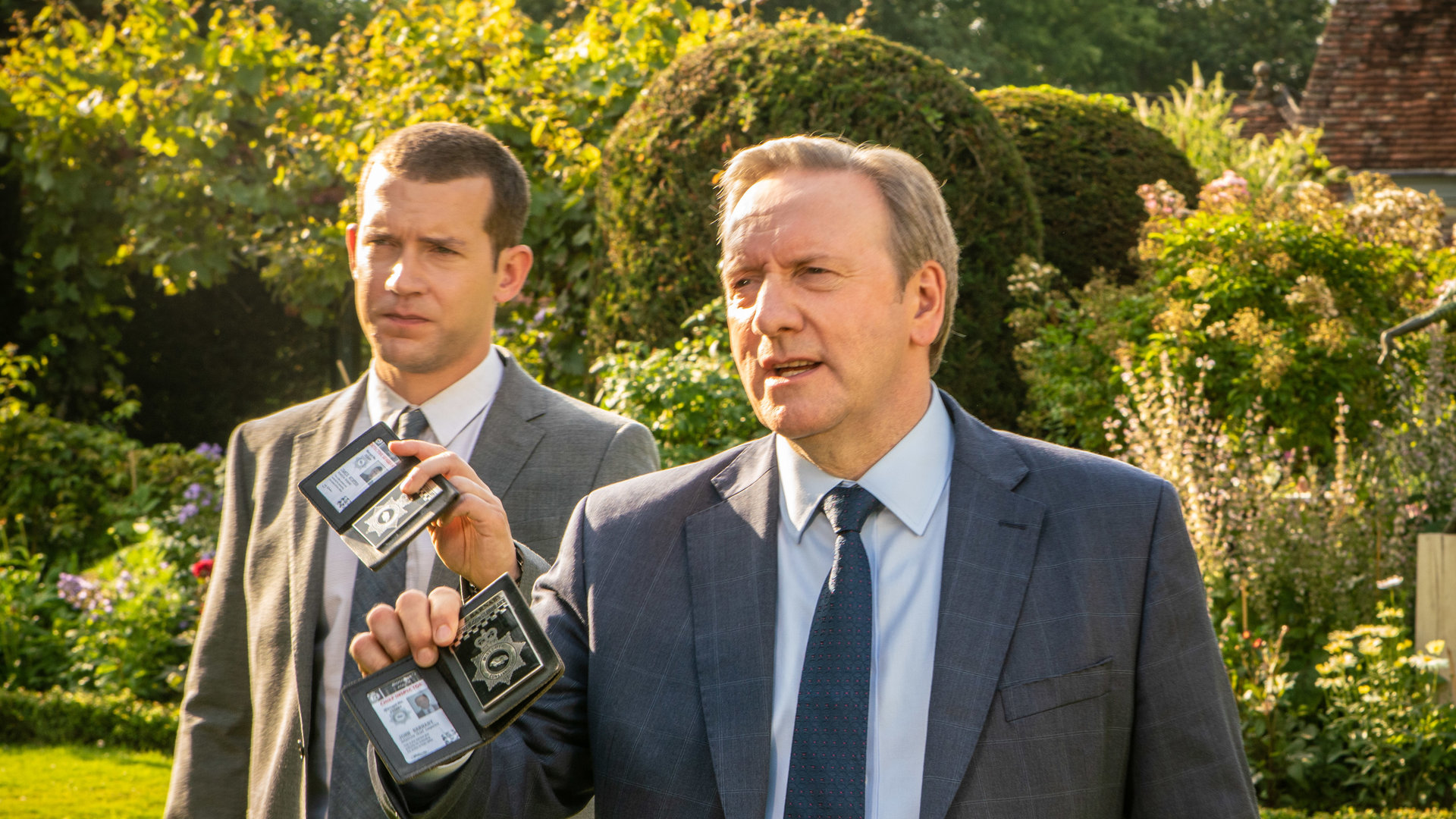 "Inspector Barnaby - Der Stachel des Todes": DCI John Barnaby (Neil Dudgeon) und DS Jamie Winter (Nick Hendrix) gehen nebeneinander über einen gepflegten Rasen. Sie halten ihrer Dienstmarken hoch, um sich als Polizisten zu erkennen zu geben.