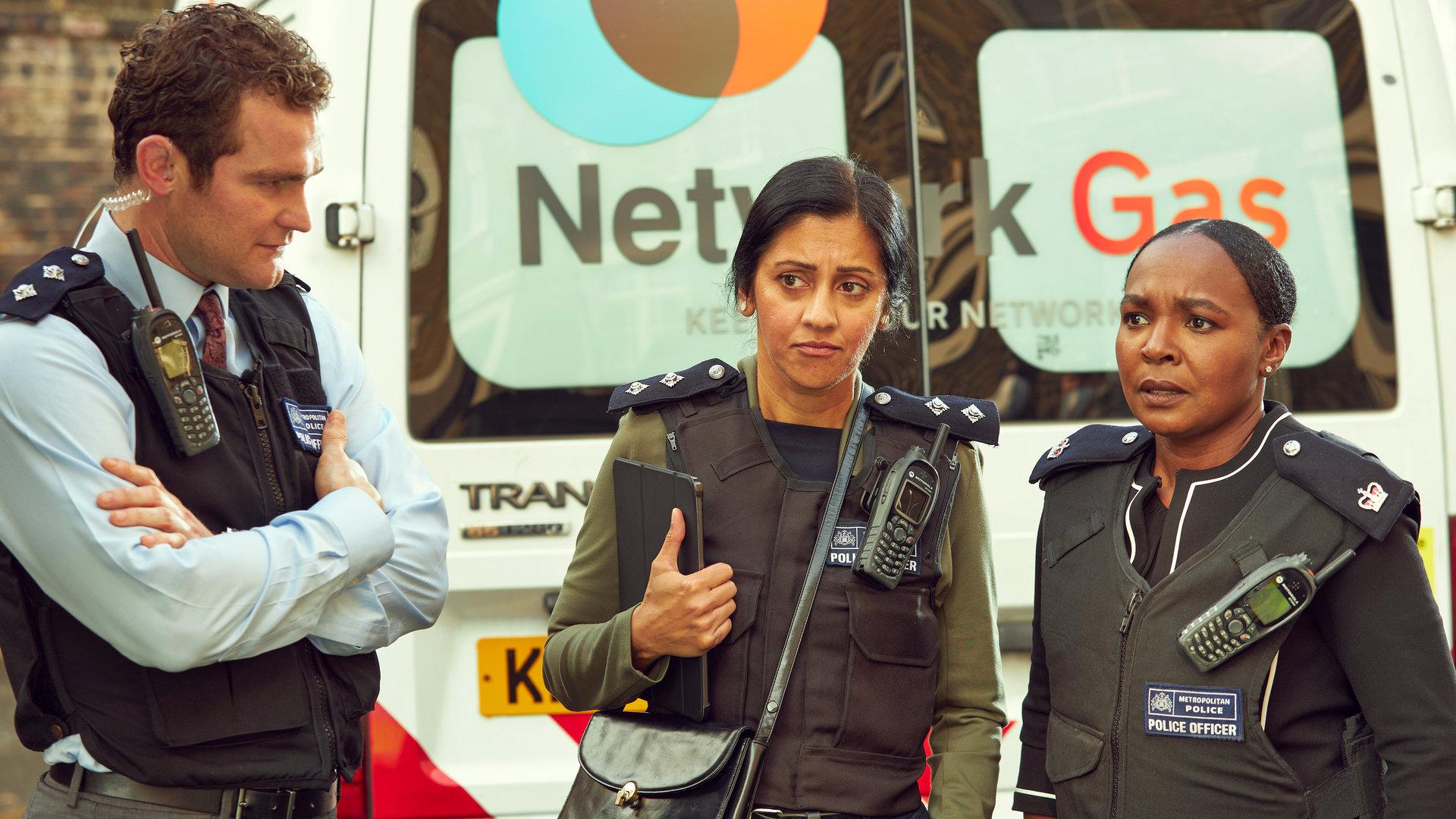 "Trigger Point: Endspiel": Thom (Mark Stanley), Desai (Manjinder Virk) und Einsatzleiterin Hamilton (Nadine Marshall) stehen mit Schutzwesten hinter einem Lieferwagen zusammen. Thom hat die Arme vor sich verschränkt und schaut die beiden Frauen an.