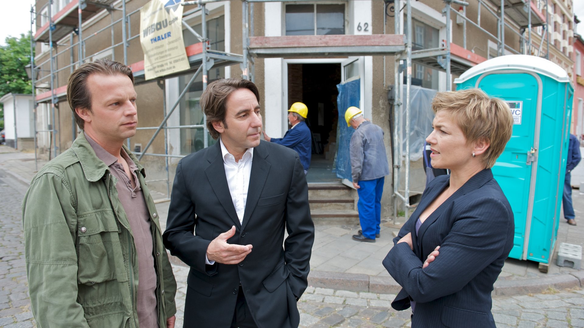 "SOKO Wismar - Das verschlossene Zimmer": Die Kommissare Katrin Börensen (Claudia Schmutzler) und Sven Herzog (Michael Härle) stehen mit Michael Borgmann (Dieter Landuris) vor dem Altstadthaus, dass Michael Borgmann im Auftrag von Thaler durchgeführt hat. Das Haus ist eingerüstet und rechts steht ein Dixi-Klo.