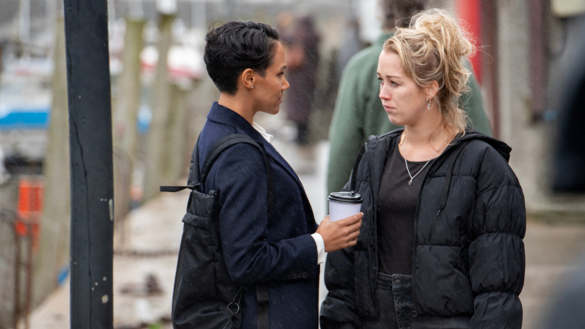 "Beyond Paradise - Der Ring": Hayley Collins (Hannah Traylen) und Kate Nolan (Lily Frazer) stehen sich auf der Straße gegenüber und schauen sich an. Beide halten Kaffeebecher in der Hand.