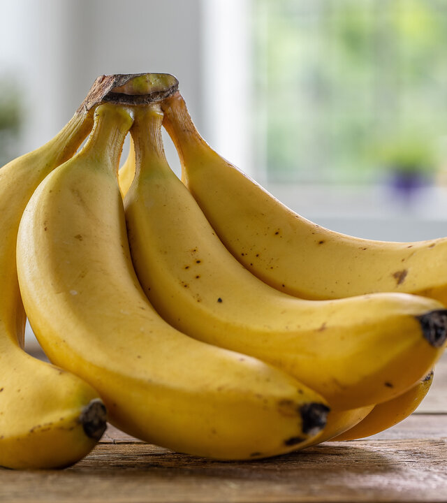 "Bananen - Genuss mit Beigeschmack": Ein Bund mit vier Bananen liegt auf einer hölzernen Arbeitsplatte.