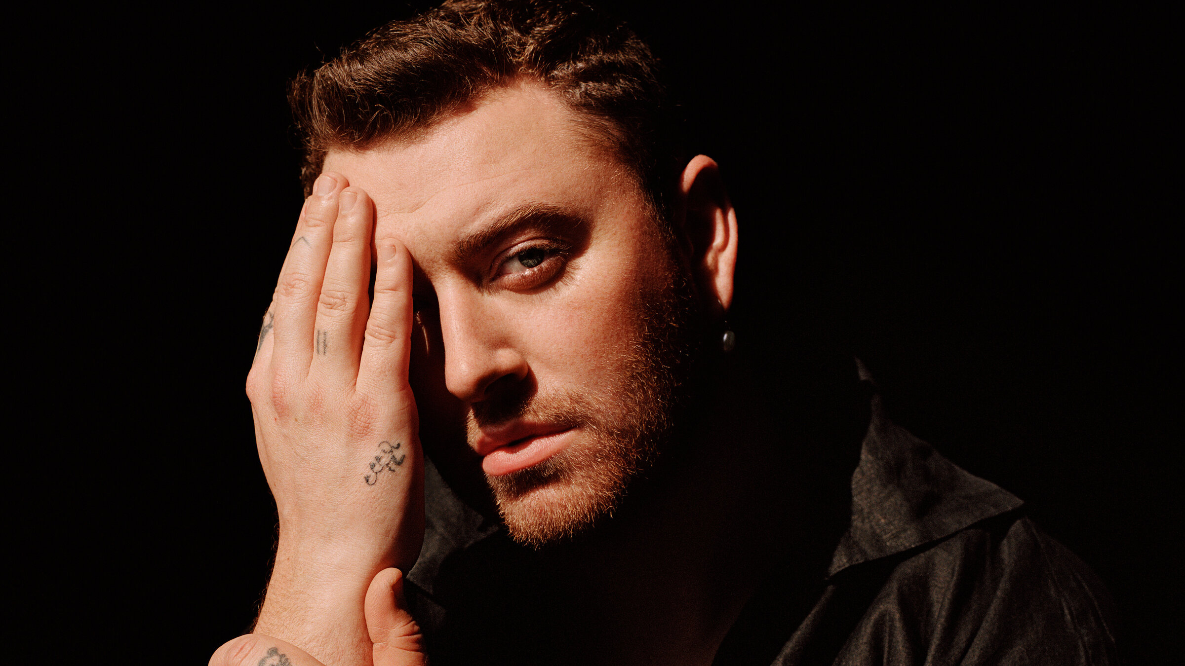 "Pop Around the Clock: Sam Smith: Live at the Royal Albert Hall": Ein Mann mit kurzen dunklen Haaren sitzt vor einem schwarzen Hintergrund mit einem schwarzen Hemd. Einen Arm hat er aufgestützt, mit der anderen Hand bedeckt er die Hälfte seines Gesichts.