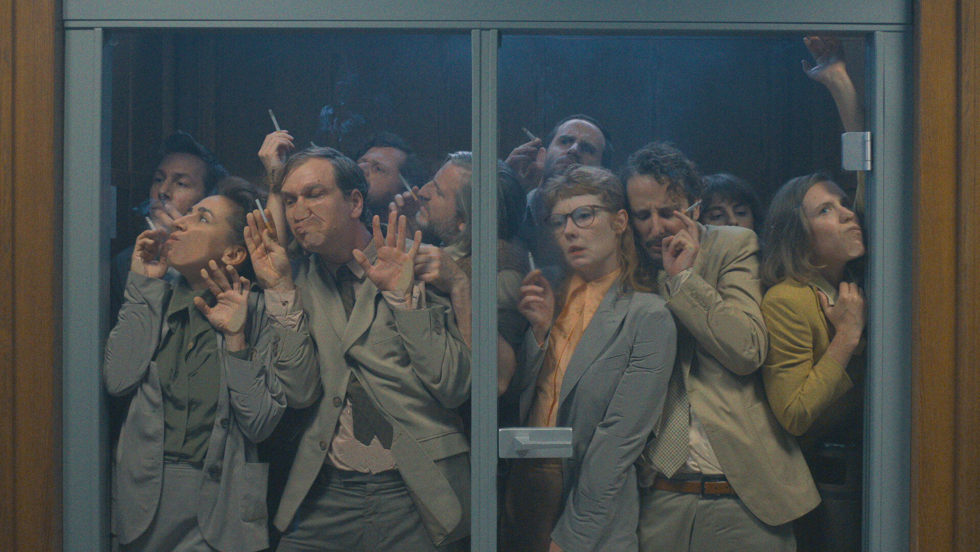 Das Manko - Eskalation in Beige: Viele Personen drängen sich in der Rauchkabine und sind durch eine Glaswand zu erkennen: Akkurat Manko (Florian Anderer), Horror Manko (Carol Schuler), Romantik Manko (Jan Krauter), Wichtig Manko (David Simon), Dumm Manko (Jonas Hien), Perplex Manko (Sebastian Grünewald), Panik Manko (Sarah Bauerett), Gute Laune Manko (Bastian Reiber), ehrgeizige Mitarbeiterin (Amelie Willberg) und Bienchen Manko (Annika Meier))