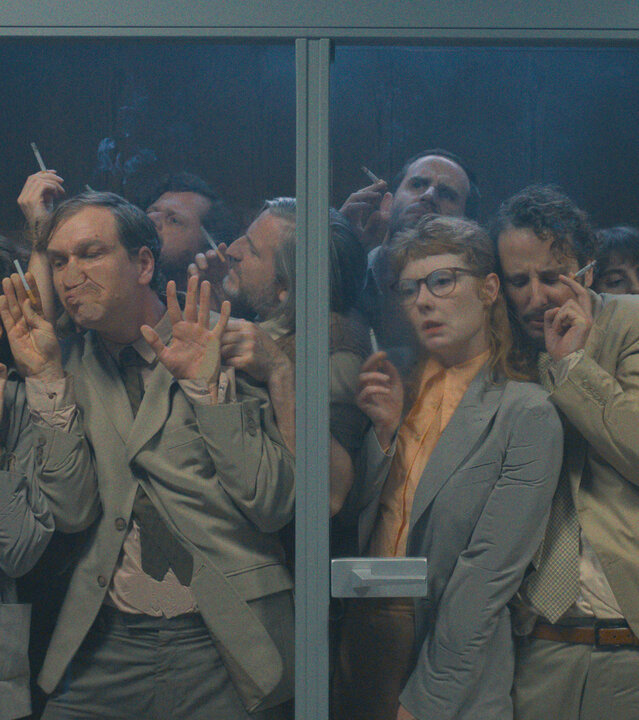 Das Manko - Eskalation in Beige: Viele Personen drängen sich in der Rauchkabine und sind durch eine Glaswand zu erkennen: Akkurat Manko (Florian Anderer), Horror Manko (Carol Schuler), Romantik Manko (Jan Krauter), Wichtig Manko (David Simon), Dumm Manko (Jonas Hien), Perplex Manko (Sebastian Grünewald), Panik Manko (Sarah Bauerett), Gute Laune Manko (Bastian Reiber), ehrgeizige Mitarbeiterin (Amelie Willberg) und Bienchen Manko (Annika Meier))