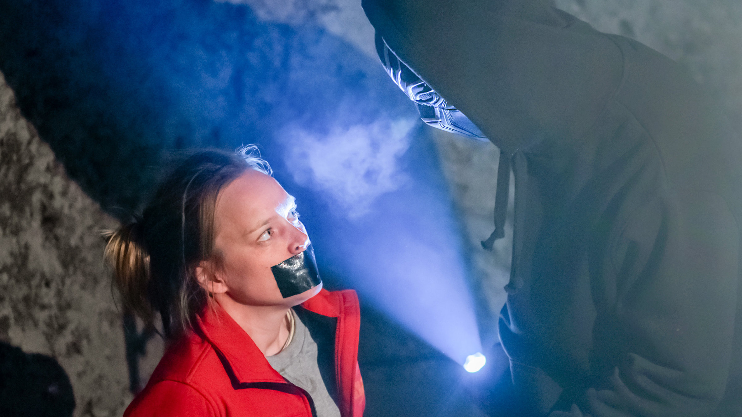 "SOKO Kitzbühel - Gefangen": In einer Höhle steht Inga Stegemann (Livia Matthes) dem maskierten Entführer (Tobias Resch) gegenüber und blickt zu ihm hinauf. An ihrem Mund ist ein schwarzes Klebeband befestigt. Der Maskierte trägt einen schwarzen Kapuzenpullover und leuchtet mit einer Taschenlampe zwischen sich und Inga Stegemann.