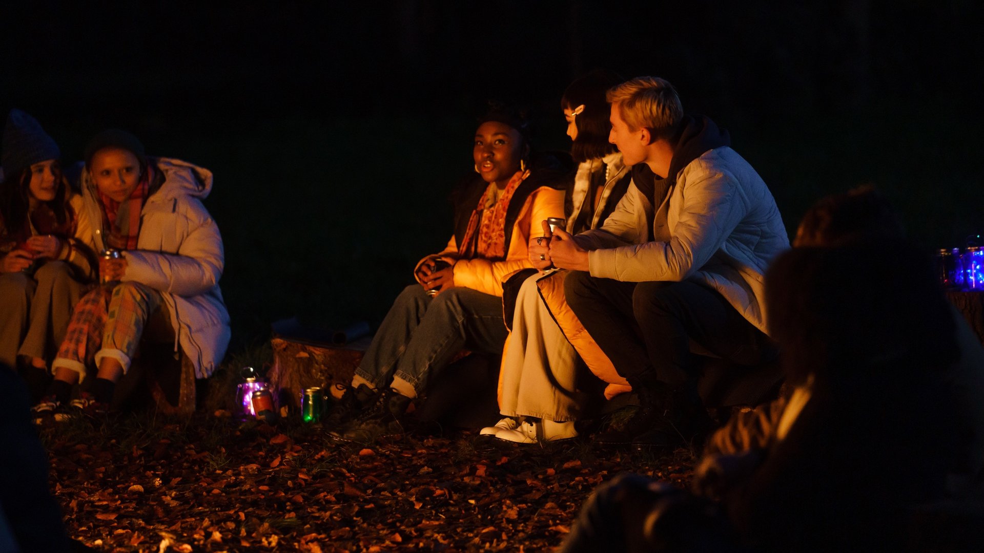 "Tell me everything: Therapie": Neve (Lauryn Ajufo), Mei (Callina Liang) und Jonny (Eden H. Davies) sitzen im Dunkeln um ein Lagerfeuer herum und unterhalten sich.