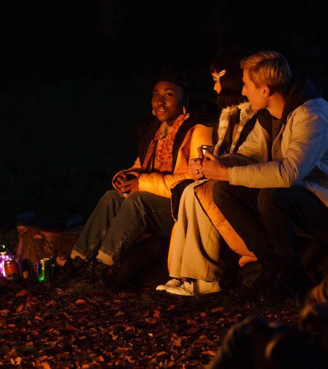 "Tell me everything: Therapie": Neve (Lauryn Ajufo), Mei (Callina Liang) und Jonny (Eden H. Davies) sitzen im Dunkeln um ein Lagerfeuer herum und unterhalten sich.