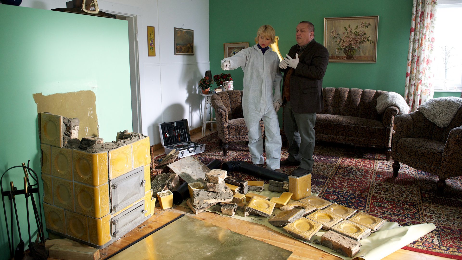 "SOKO Wismar - Über den Dächern von Wismar": Roswitha Prinzler (Silke Matthias) und Jan Reuter (Udo Kroschwald) stehen in einem Wohnzimmer. Im Hintergrund sind eine Sitzecke mit einer Couch und zwei Sesseln zu sehen. Roswitha, gekleidet in einen weißen Schutzanzug, zeigt auf einen auseinander gebauten Ofen mitten im Raum.