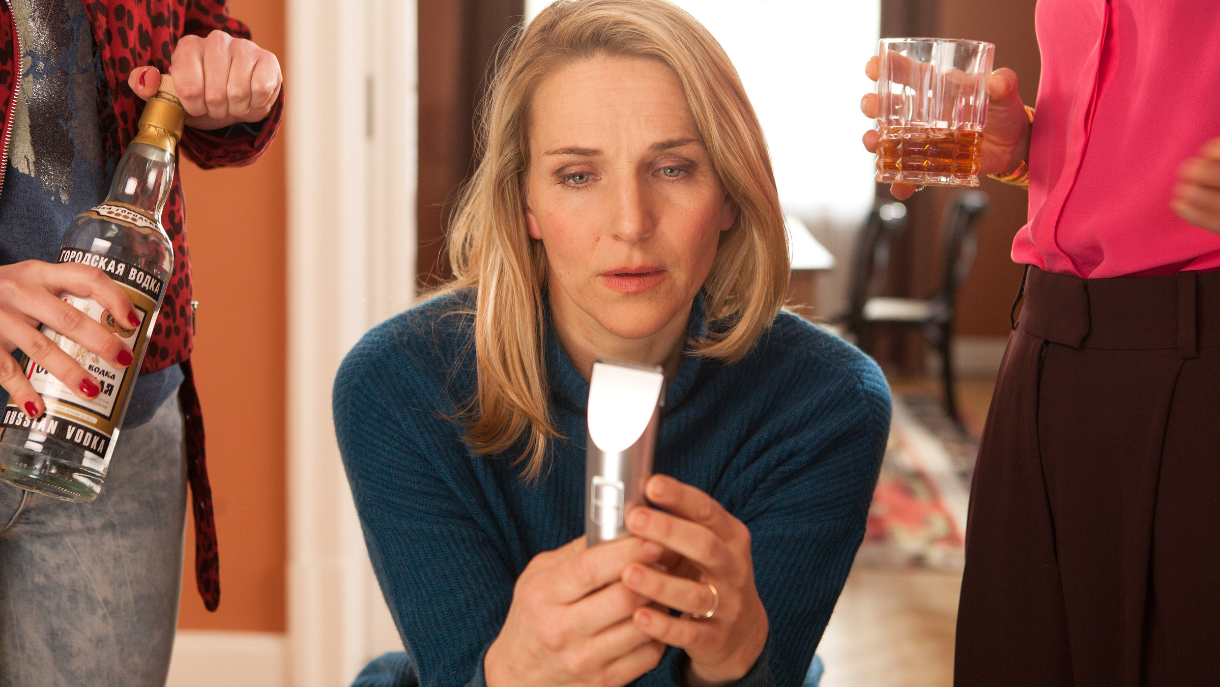 "Fritzie – Der Himmel muss warten – Kurswechsel": Fritzie (Tanja Wedhorn) sitzt in ihrem Wohnzimmer und starrt auf einen elektrischen Rasierer, den sie in der Hand hält. Links neben ihr sind Melanies (Anna Herrmann) Hände zu sehen, die eine Wodkaflasche halten. Rechts von Fritzie ist in Selmas (Neda Rahmanian) Hand ein Glas mit einer klarbraunen Flüssigkeit zu sehen.