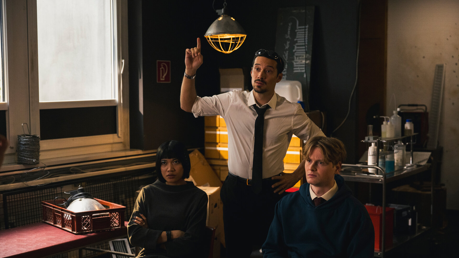 "SPYON - Die goldenen Regeln": In einem Innenraum: Yuki (Nhung Hong) und Asel Asanow (Lukas Zumbrock) sitzen nebeneinander und blicken seitlich nach vorne. Andi (Klaus Steinbacher) steht zwischen den beiden und hebt seine Hand mit ausgestrecktem Finger. Hinter ihnen befindet sich ein Fenster, daneben hängt eine Leuchte vor einem Schrank. Neben dem Schrank steht ein Servierwagen mit diversen Utensilien.