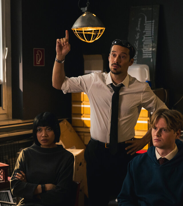 "SPYON - Die goldenen Regeln": In einem Innenraum: Yuki (Nhung Hong) und Asel Asanow (Lukas Zumbrock) sitzen nebeneinander und blicken seitlich nach vorne. Andi (Klaus Steinbacher) steht zwischen den beiden und hebt seine Hand mit ausgestrecktem Finger. Hinter ihnen befindet sich ein Fenster, daneben hängt eine Leuchte vor einem Schrank. Neben dem Schrank steht ein Servierwagen mit diversen Utensilien.