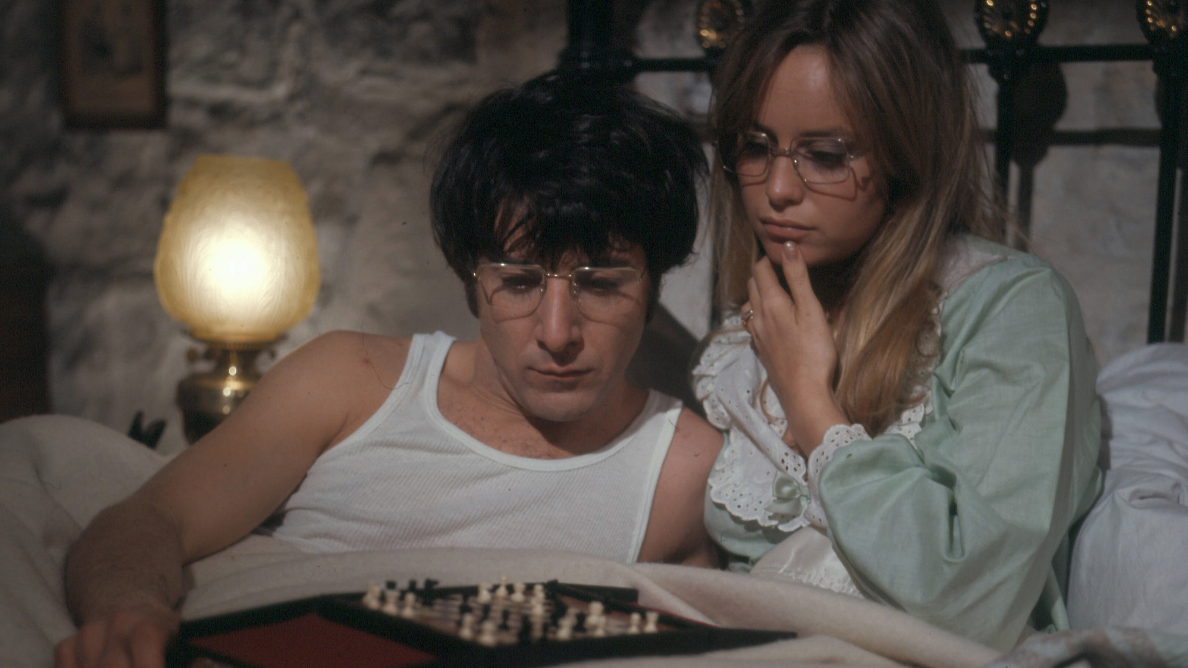 "Wer Gewalt sät": Ein junger Mann und eine Frau, beide mit Brille, liegen und sitzen in Unterhemd beziehungsweise Pyjama im Bett und grübeln über einer Partie Schach. Im Hintergrund erleuchtet eine Stehlampe das Zimmer.