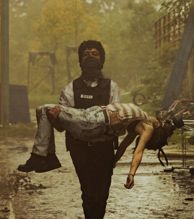 "Phoenix - Change Or Die: Wem gehört die Welt?": Polizist Amin Kacem (Mustapha Abourachid), ausgestattet mit einer schusssicheren Weste und mit einem Tuch vor dem Mund, trägt eine leblos wirkende Eloïse (Marie Colomb) in seinen Armen durch die rauchgeschwängerte Luft.