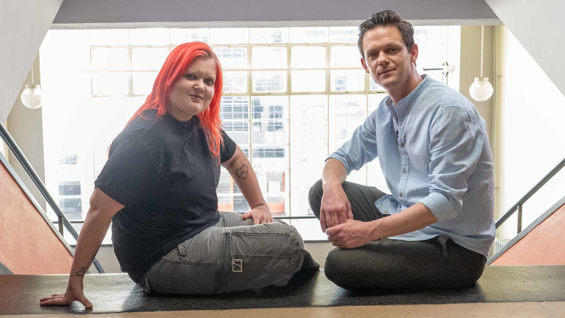 "Pop Around @ Bauhaus: Alma": Sängerin Alma mit rot-gefärbten langen Haaren und schwarzer Kleidung sitzt gegenüber im Schneidersitz von Moderator Jo Schück, der ein hellblaues Hemd und Jeans trägt.