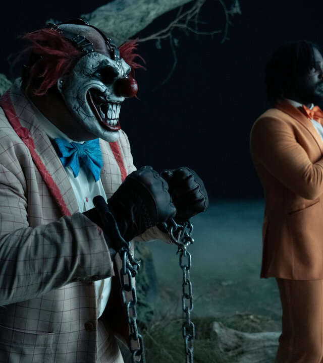 "Twisted Metal - M4YH3M": Sweet Tooth (Joe Seanoa) steht bei Nacht im Freien vor einem Baum. Er trägt einen Anzug und eine breit grinsende, bereits etwas mitgenommene Clownsmaske mit seitlich abstehenden roten Haaren, die sein Gesicht vollständig bedeckt. Seine Hände sind mit einer langen silbernen Kette gefesselt. Im Hintergrund stehen Mike (Tahj Vaughans) und Stu (Mike Mitchell) nebeneinander, beide blicken verängstigt nach vorne.