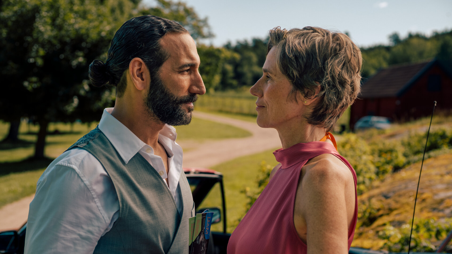 "Inga Lindström: Rezept für die Liebe": Frederika (Julia Bremermann) und Kian (Massimo Sinato) stehen im Freien dicht beieinander und sehen sich an. Im Hintergrund liegt eine sich schlängelnde Schotterstraße, umgeben von Bäumen und Wiese. Direkt hinter dem Paar steht ein rotes Cabrio mit offenem Dach.