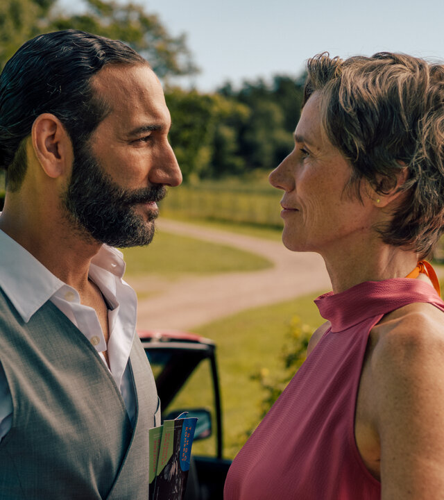 "Inga Lindström: Rezept für die Liebe": Frederika (Julia Bremermann) und Kian (Massimo Sinato) stehen im Freien dicht beieinander und sehen sich an. Im Hintergrund liegt eine sich schlängelnde Schotterstraße, umgeben von Bäumen und Wiese. Direkt hinter dem Paar steht ein rotes Cabrio mit offenem Dach.