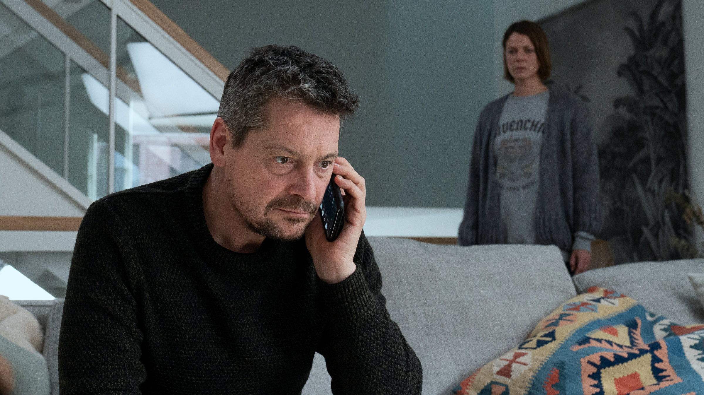 "Alles auf Rot": Kessel (Fritz Karl) sitzt in einem Wohnzimmer auf einem grauen Sofa und telefoniert. Claire (Jessica Schwarz) steht schräg hinter ihm an einer Treppe und belauscht das Telefonat.