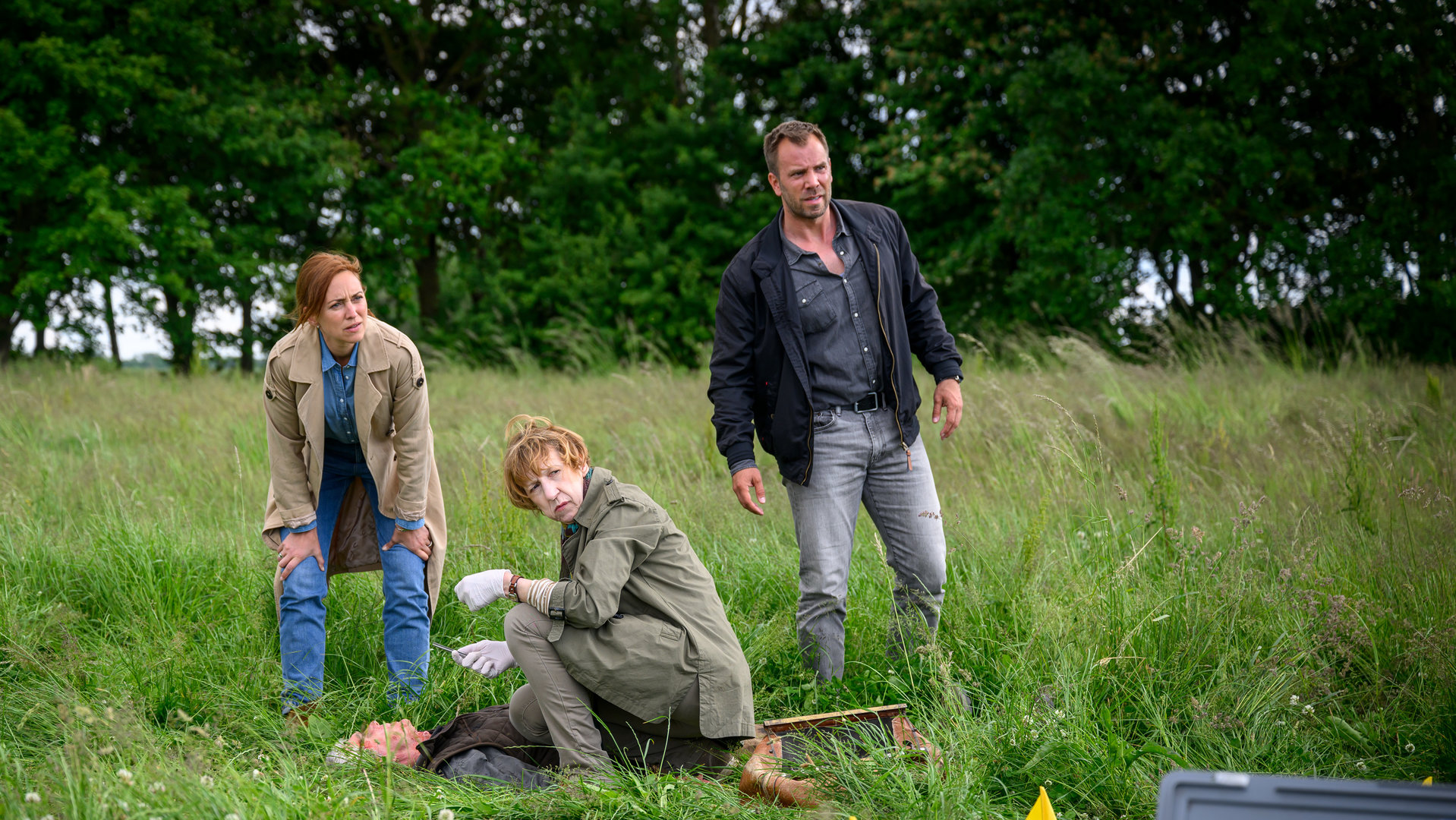 "SOKO Wismar – Der Umbetter": Auf einer Wiese umgeben von Bäumen, liegt das Opfer Enno Kämpfer (Carsten Voigt) auf dem Boden. Daneben kniet Rechtsmedizinerin Dr. Helene Sturbeck (Katharina Blaschke), begleitet von Kommissarin Karoline Joost (Nike Fuhrmann) und Kommissar Lars Pöhlmann (Dominic Boeer).