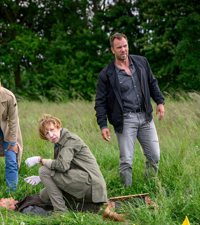 "SOKO Wismar – Der Umbetter": Auf einer Wiese umgeben von Bäumen, liegt das Opfer Enno Kämpfer (Carsten Voigt) auf dem Boden. Daneben kniet Rechtsmedizinerin Dr. Helene Sturbeck (Katharina Blaschke), begleitet von Kommissarin Karoline Joost (Nike Fuhrmann) und Kommissar Lars Pöhlmann (Dominic Boeer).