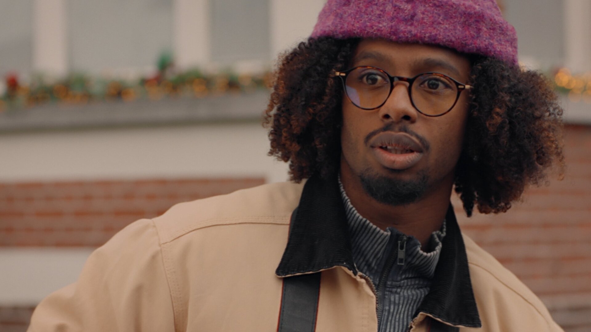 "Single Bells - 12 Dates till Christmas: Anthony": Anthony (Malik Mohammed) in Nahaufnahme. Er trägt eine Brille und einen gepflegten Kinn- und Oberlippenbart. Zahlreiche gelockte Haare schauen unter seiner lilafarbenen Wollmütze hervor.