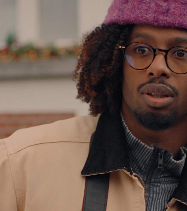 "Single Bells - 12 Dates till Christmas: Anthony": Anthony (Malik Mohammed) in Nahaufnahme. Er trägt eine Brille und einen gepflegten Kinn- und Oberlippenbart. Zahlreiche gelockte Haare schauen unter seiner lilafarbenen Wollmütze hervor.