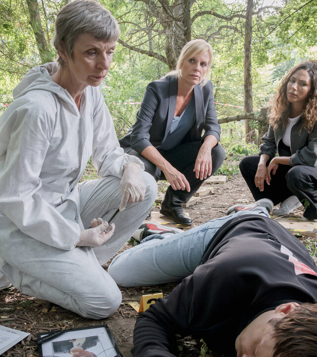 "SOKO Leipzig - Nur ein Foto": Prof. Dr. Rossi (Anna Stieblich) kniet mit Ina (Melanie Marschke) und Kim (Amy Mußul)  am Tatort in einem Waldfriedhof. Im Vordergrund liegt eine männliche Leiche (Paul Maximilian Pira).