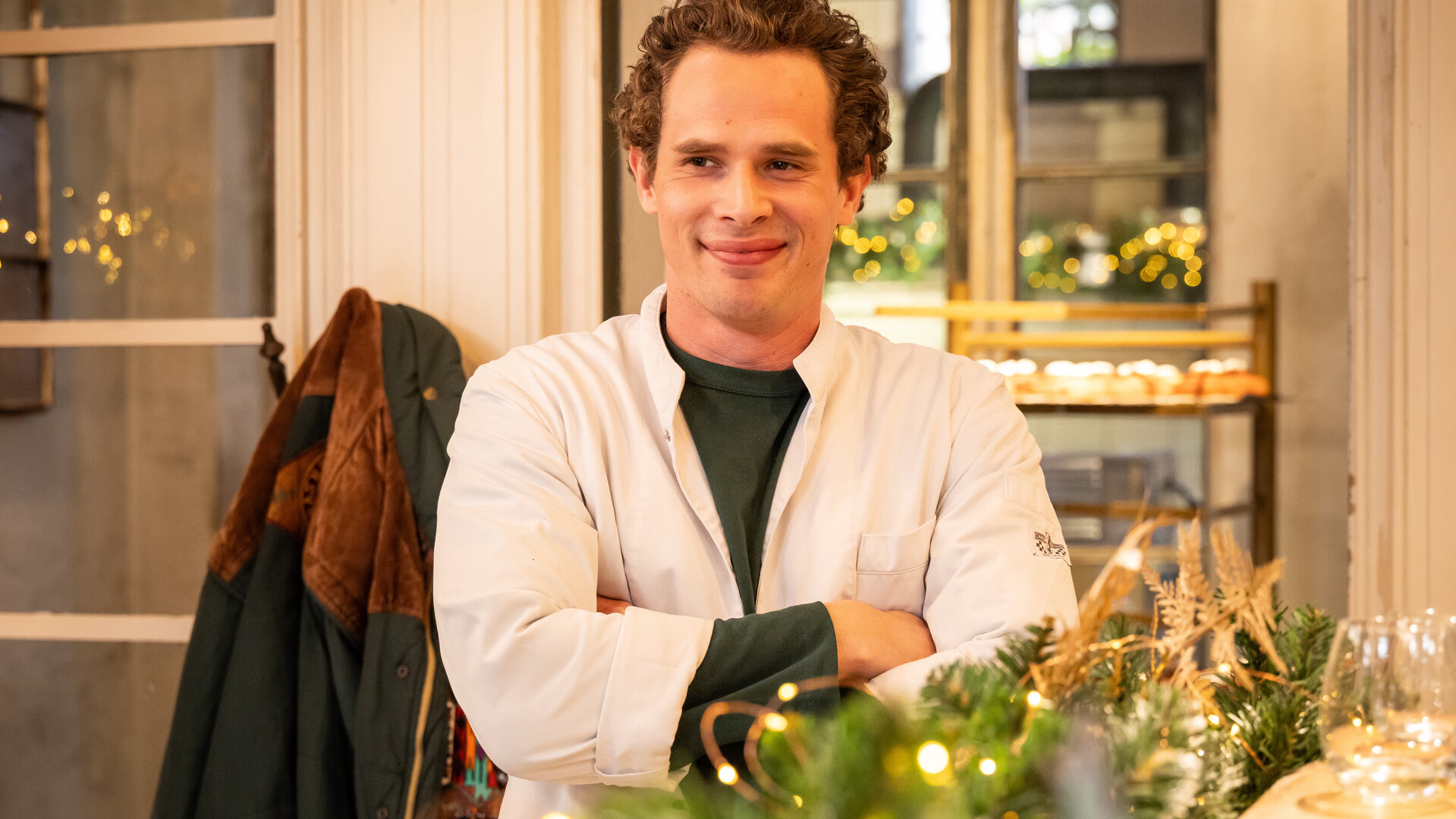 "Single Bells - Only Losers for Annie? - Diane": Jens (Flor Van Severen) steht mit verschränkten Armen und einem breiten Lächeln in seiner Bäckerei.