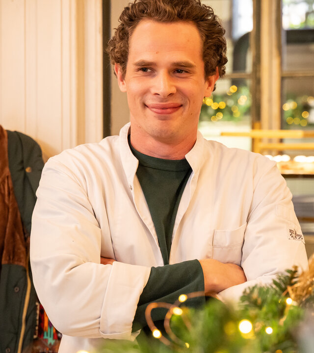 "Single Bells - Only Losers for Annie? - Diane": Jens (Flor Van Severen) steht mit verschränkten Armen und einem breiten Lächeln in seiner Bäckerei.
