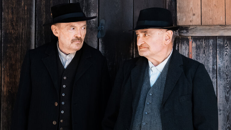 "Sturm kommt auf": Schuster Julius Kraus (Josef Hader) und Silvan Heingeiger (Sigi Zimmerschied) stehen vor einer Holzwand nebeneinander und schauen sich an.