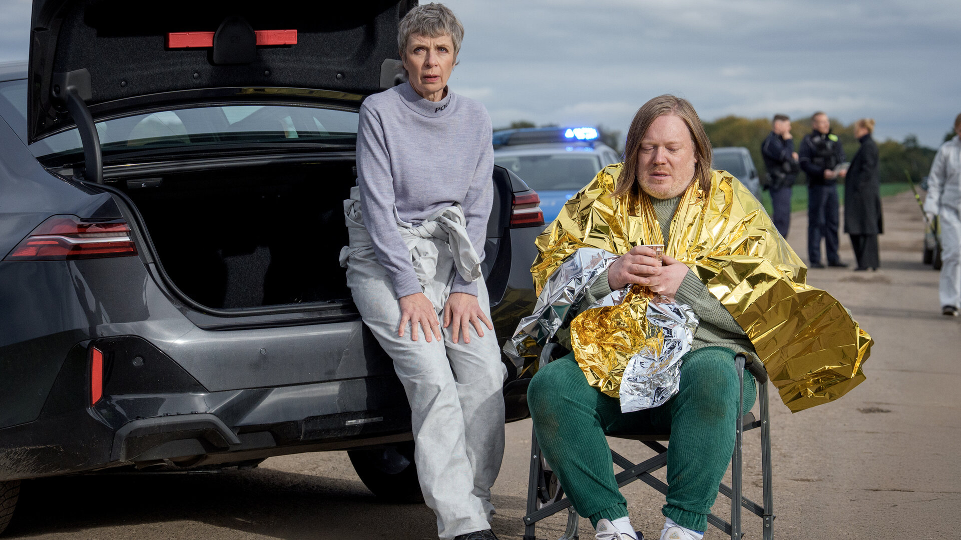 "SOKO Leipzig - Heute sterbe ich nicht": Prof. Sabine Rossi (Anna Stieblich) sitzt an den offenen Kofferraum eines Autos gelehnt neben Lars Roth (Christoph Franken), der in eine Rettungsdecke gehüllt in einem Klappstuhl sitzt. Er blickt betrübt auf den Boden. Im Hintergrund in der Unschärfe stehen weitere Polizisten neben weiteren Fahrzeugen.