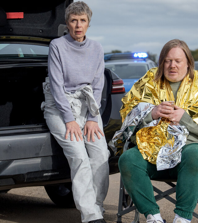 "SOKO Leipzig - Heute sterbe ich nicht": Prof. Sabine Rossi (Anna Stieblich) sitzt an den offenen Kofferraum eines Autos gelehnt neben Lars Roth (Christoph Franken), der in eine Rettungsdecke gehüllt in einem Klappstuhl sitzt. Er blickt betrübt auf den Boden. Im Hintergrund in der Unschärfe stehen weitere Polizisten neben weiteren Fahrzeugen.