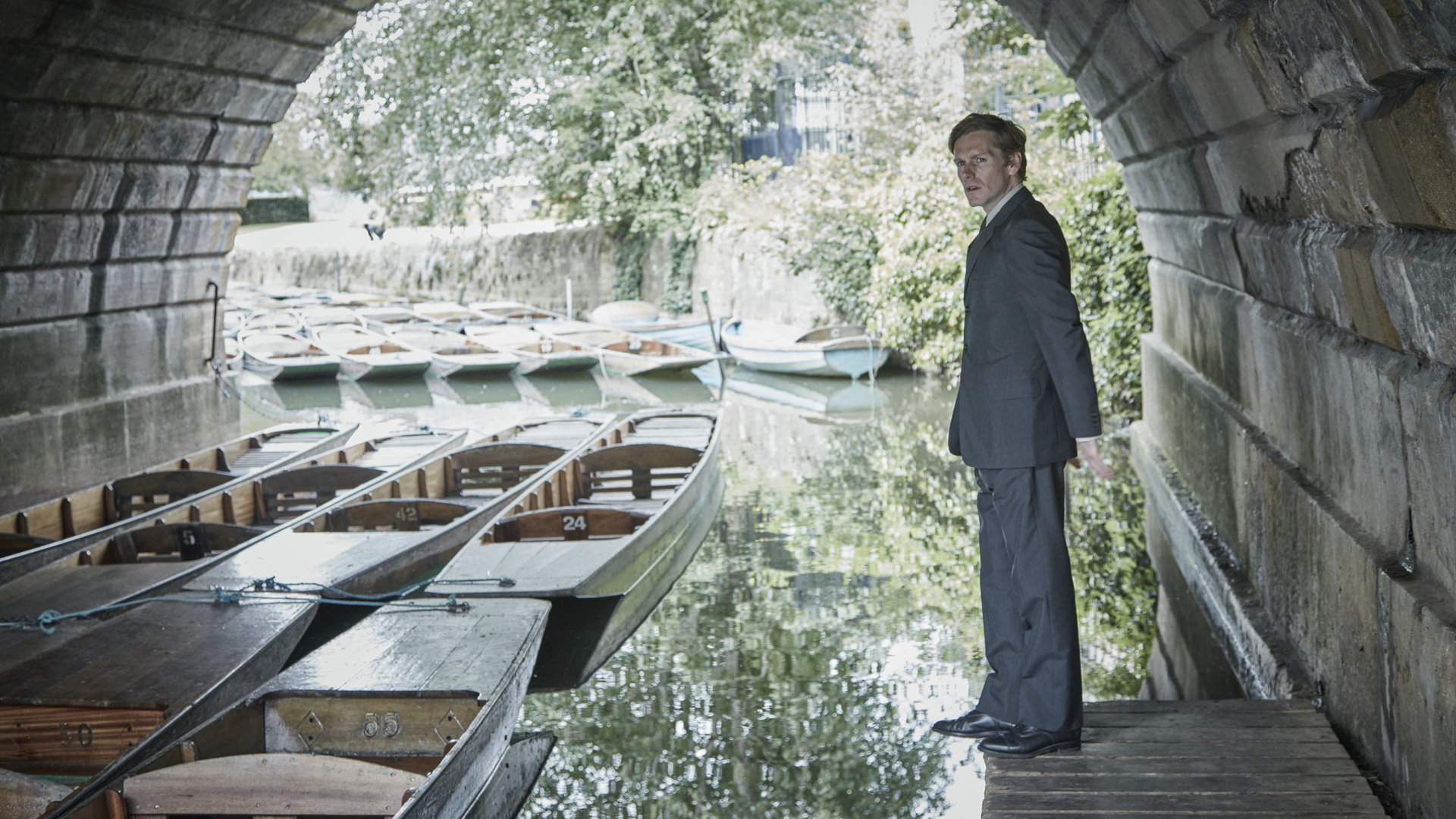 "Der junge Inspektor Morse - Totenmasken": Endeavour Morse (Shaun Evans) steht am Rande eines Bootsanlege-Platzes unter einer Brücke und schaut sich um. In seiner Nähe liegen viele kleine Holzboote im Wasser.
