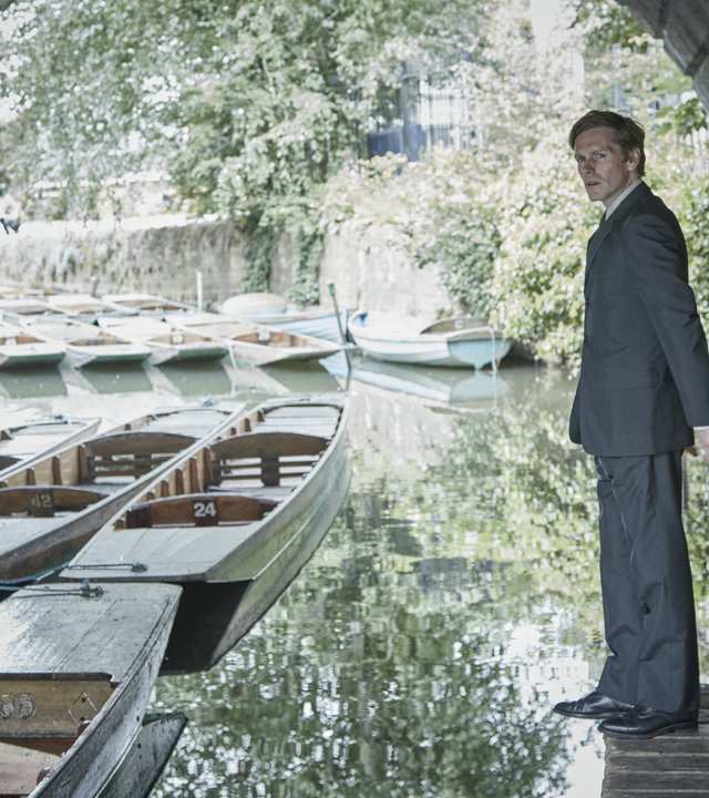 "Der junge Inspektor Morse - Totenmasken": Endeavour Morse (Shaun Evans) steht am Rande eines Bootsanlege-Platzes unter einer Brücke und schaut sich um. In seiner Nähe liegen viele kleine Holzboote im Wasser.