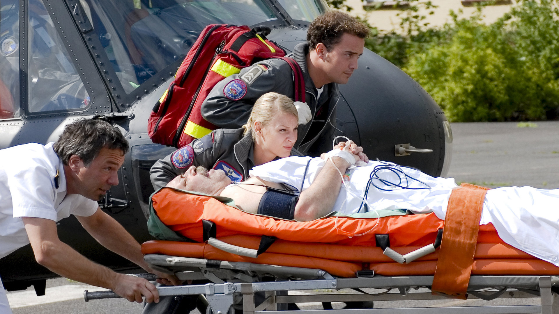 "Die Rettungsflieger - Muttersorgen": Dr. Sabine Petersen (Marlene Marlow) und Johannes von Storkow (Patrick Wolf) neben einer Krankentrage mit dem verletzten Bauern Heinrich (Klaus Dittmann), die ein Pfleger vom Hubschrauber Richtung Krankenhaus schiebt.