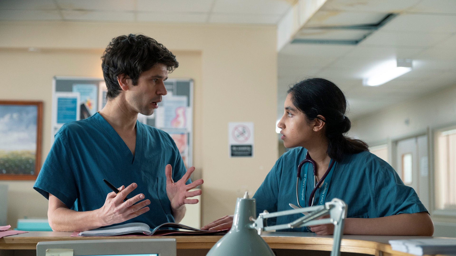 "This Is Going To Hurt - Sisyphos ist müde": Shruti (Ambika Mod) und Adam (Ben Whishaw) lehnen an der Rezeption einer Krankenstation und unterhalten sich.