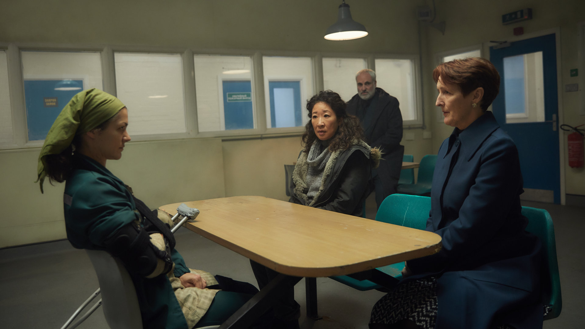 "Killing Eve - Steckt mich ins Loch": Nadja (Olivia Ross) sitzt Eve (Sandra Oh) und Carolyn Martens (Fiona Shaw) an einem Tisch im Besuchszimmer eines Gefängnisses gegenüber. Die Frauen am Tisch schauen sich ernst an, während Konstantin (Kim Bodnia) im Hintergrund das Gespräch beobachtet.