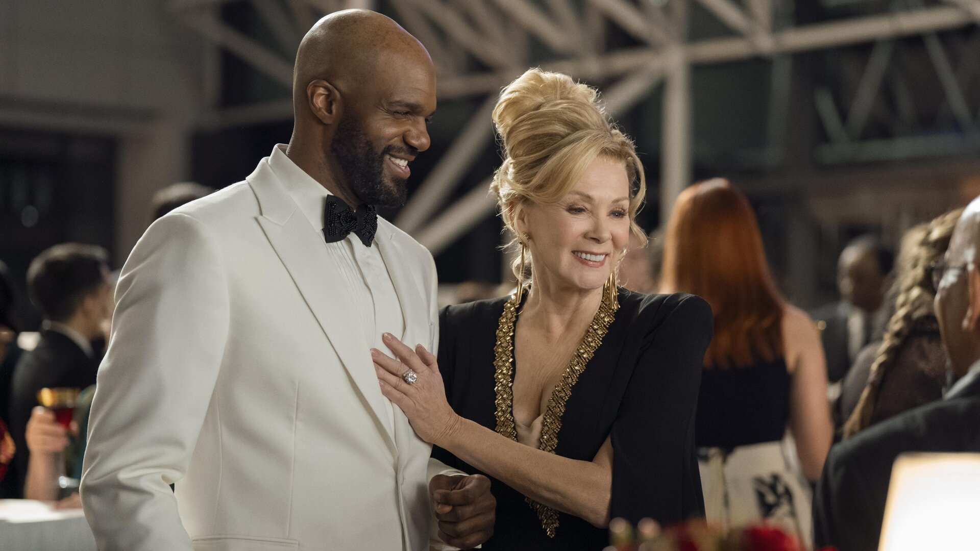 "Hacks - Verändere die Welt mit Lachern": Deborah (Jean Smart) steht im schicken Abendkleid neben dem mit weißen Smoking festlich gekleidetem Marcus (Carl Clemons-Hopkins). Deborah hat ihre Hand auf dessen Brust gelegt und beide lachen glücklich und gelöst. Im Hintergrund befinden sich weitere Personen (Komparsen).