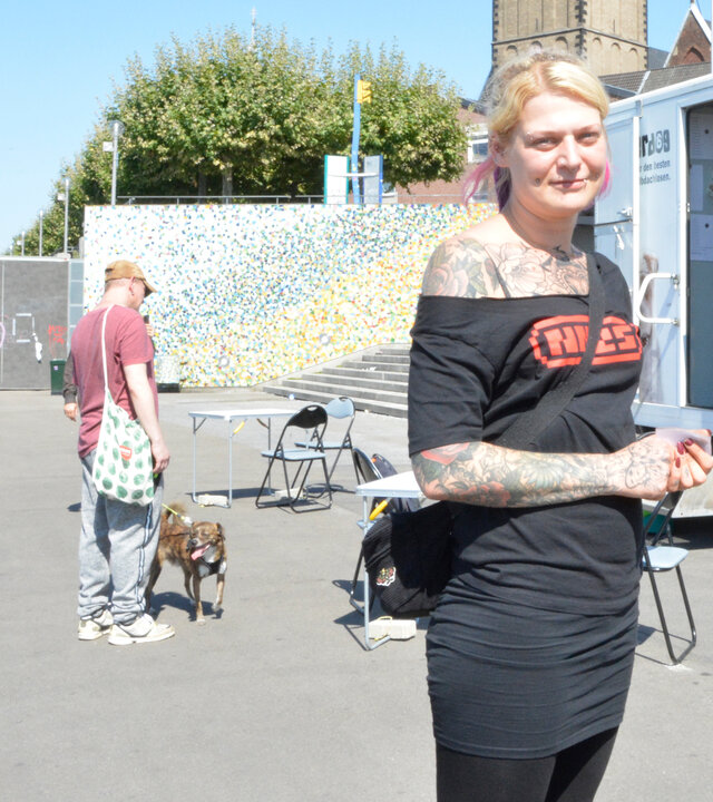 "Leben ist mehr! - Hilfe für Menschen und Hunde": An den Düsseldorfer Rheintreppen steht die Sozialarbeiterin Jana vor einem Wagen mit der Aufschrift "Underdog" und schaut lächelnd in die Kamera. Im Hintergrund stehen weitere Personen mit Hunden.