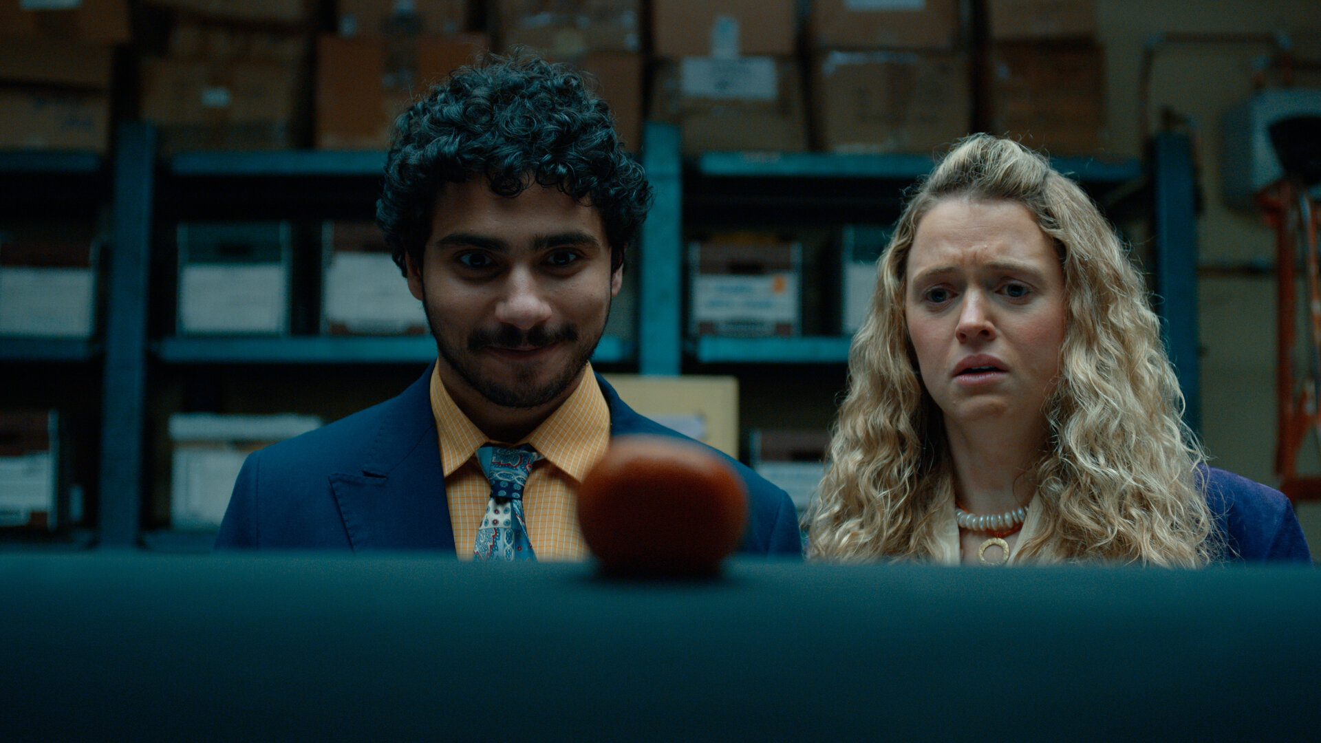 "Extraordinary - Hallo Fremder": Carrie (Sofia Oxenham) und Kash (Bilal Hasna) stehen nebeneinander in einem Lagerraum mit blauen Metallregalen und zahlreichen Kartons. Beide blicken konzentriert auf einen runden, rot-orangefarbenen Gegenstand im Vordergrund, wobei Kash zufrieden wirkt, während Carries Gesichtsausdruck deutlich besorgt erscheint.
