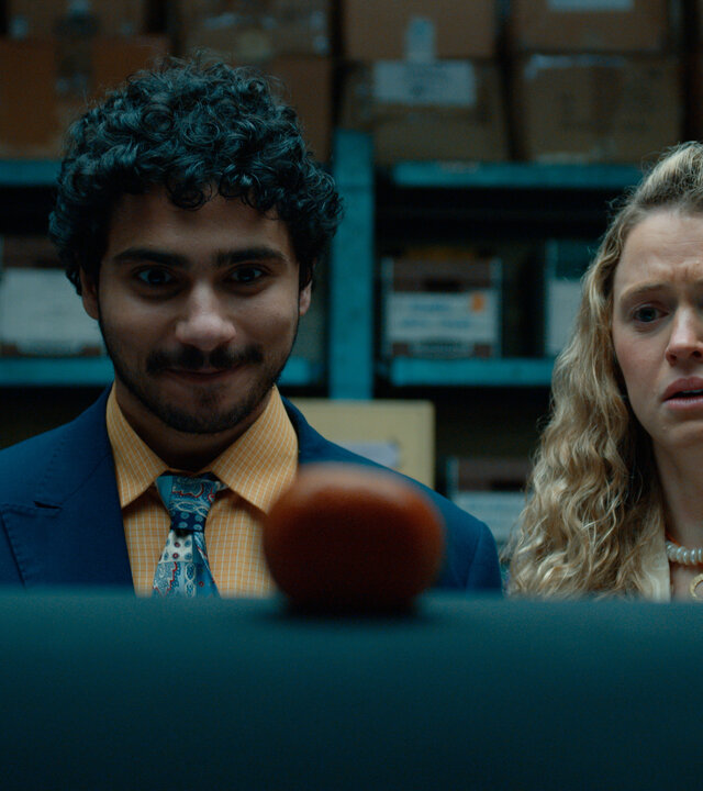 "Extraordinary - Hallo Fremder": Carrie (Sofia Oxenham) und Kash (Bilal Hasna) stehen nebeneinander in einem Lagerraum mit blauen Metallregalen und zahlreichen Kartons. Beide blicken konzentriert auf einen runden, rot-orangefarbenen Gegenstand im Vordergrund, wobei Kash zufrieden wirkt, während Carries Gesichtsausdruck deutlich besorgt erscheint.