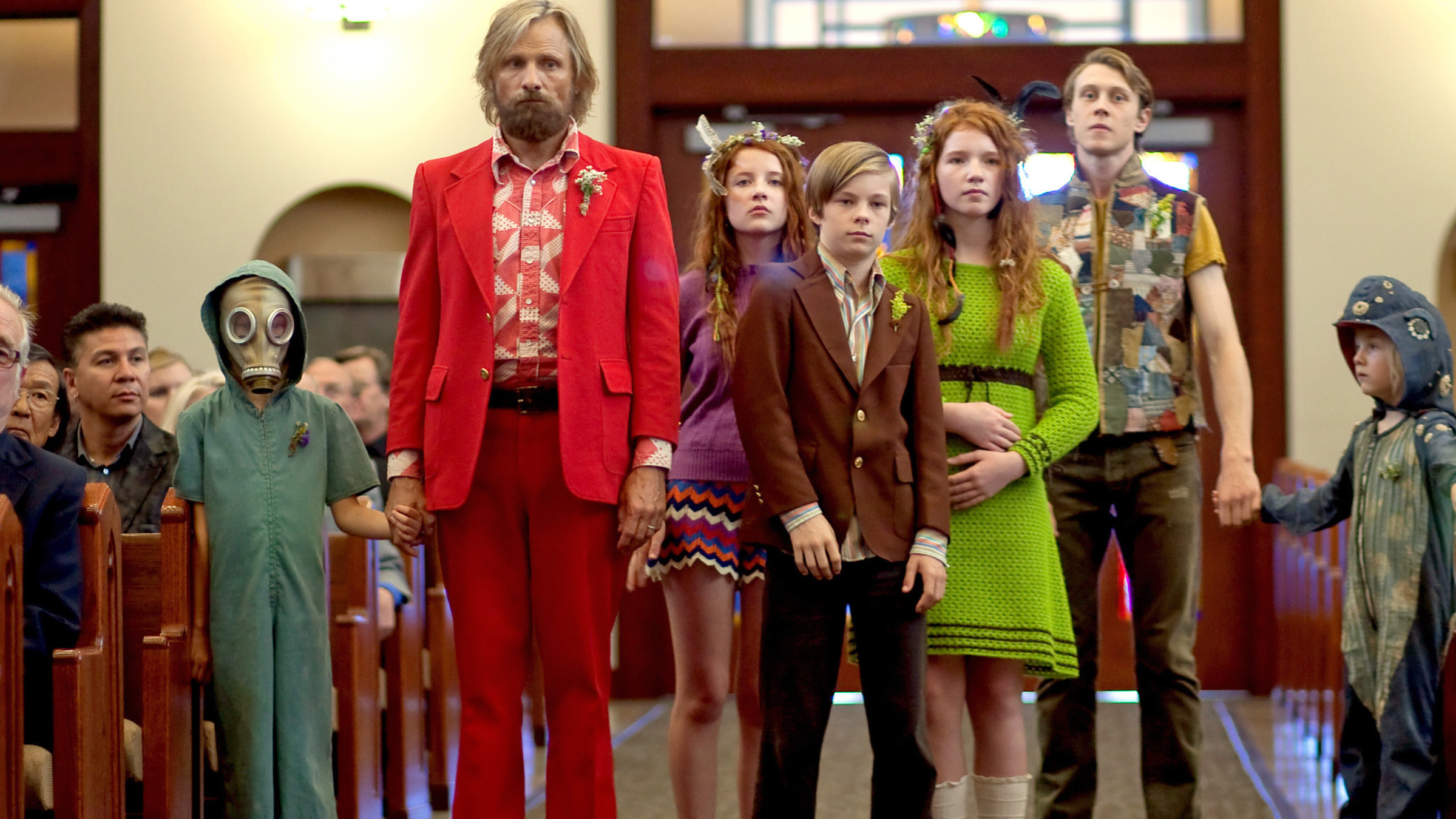 "Captain Fantastic - Einmal Wildnis und zurück": Ben (Viggo Mortensen), Nai (Charlie Shotwell), Bodevan (George MacKay), Kielyr ‎‎(Samantha Isler), Rellian (Nicholas Hamilton), Vespyr (Annalise Basso) ‎und Zaja (Shree Crooks) stehen im Mittelgang eines Kirchenraums und tragen bunte Kleidung. ‎