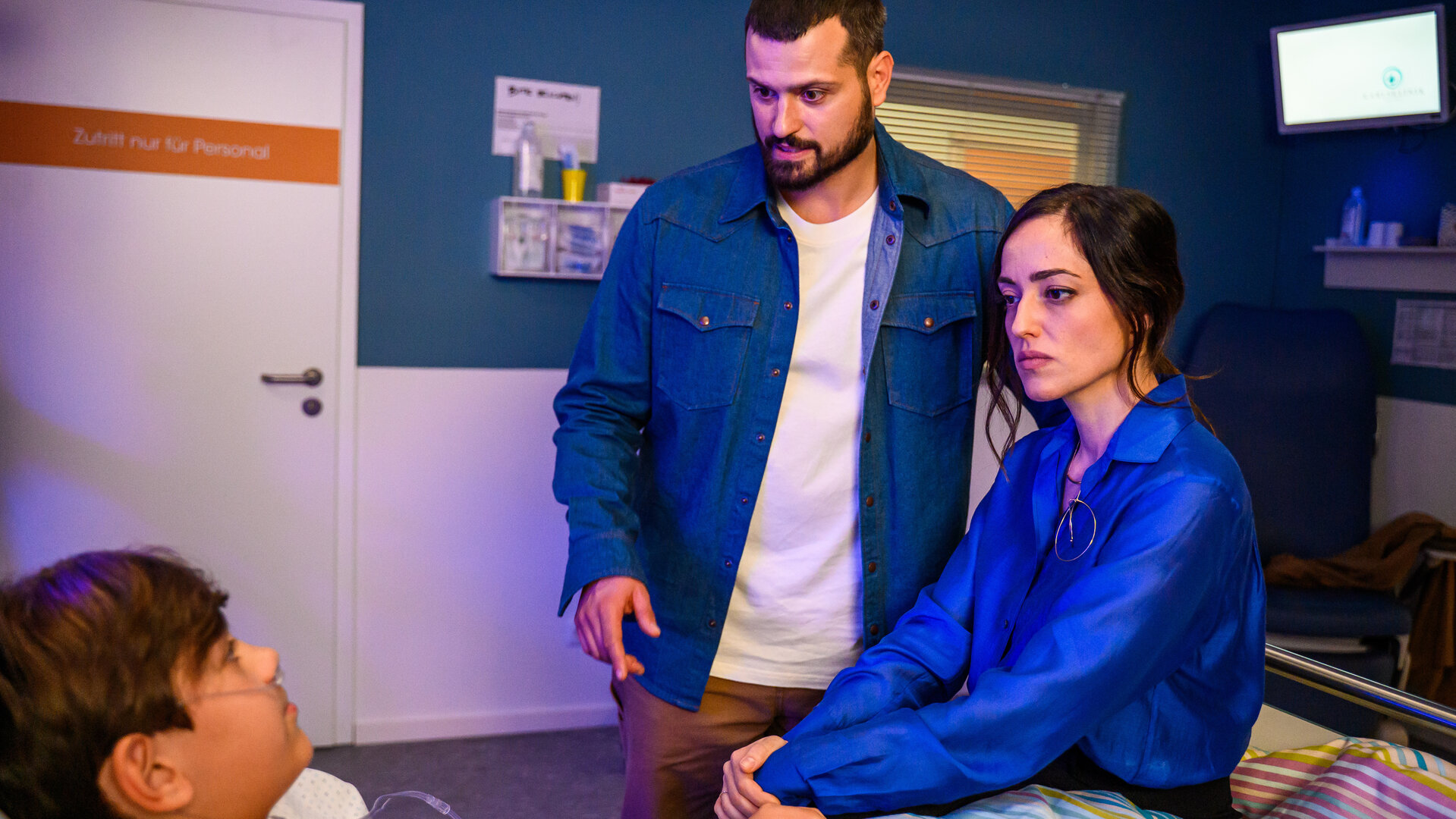 "Bettys Diagnose - Ganz der Papa": Dr. Şahin (Nicolas Fethi Türksever) und Elin Şahin (Céline Yildirim) sind am Patientenbett ihres Sohnes Nico (Caspar Boecken). Oben hängt ein Monitor an der Wand, an der Seite im Hintergrund ist eine Tür.