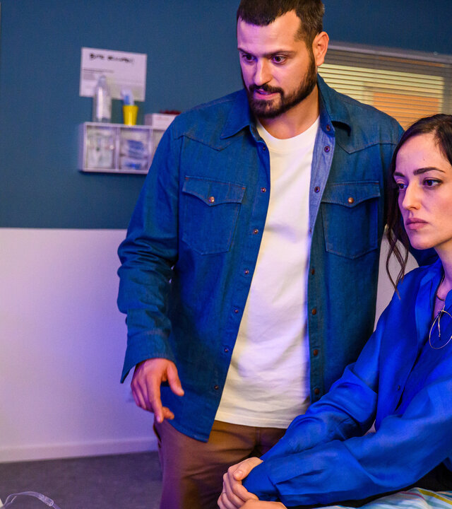 "Bettys Diagnose - Ganz der Papa": Dr. Şahin (Nicolas Fethi Türksever) und Elin Şahin (Céline Yildirim) sind am Patientenbett ihres Sohnes Nico (Caspar Boecken). Oben hängt ein Monitor an der Wand, an der Seite im Hintergrund ist eine Tür.