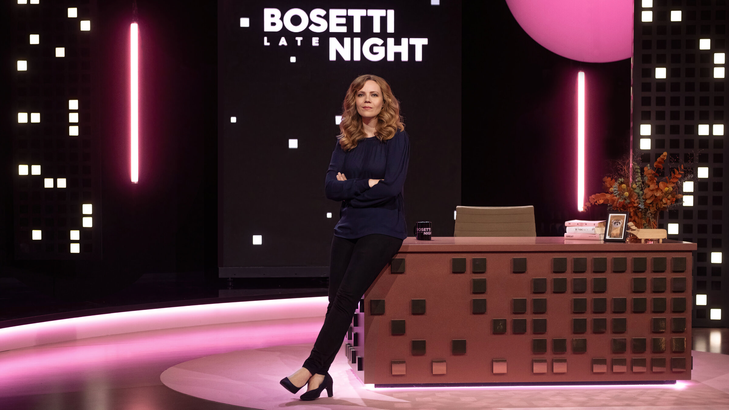 "Bosetti Late Night": Sarah Bosetti sitzt mit verschränkten Armen an der Tischkante ihres Tisches auf der Bühne.